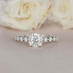 a close up view of a diamond engagement ring with white roses in the foreground