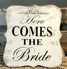 a white sign that says here comes the bride on top of a wooden bench with lace