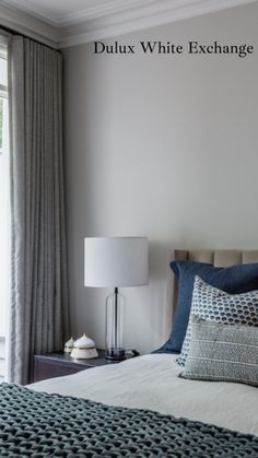 a bed with blue and white pillows in a bedroom next to a large window that reads dulux white exchangeage