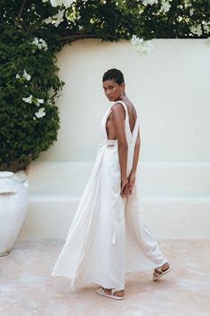 a woman in a white dress standing next to a wall with flowers growing on it