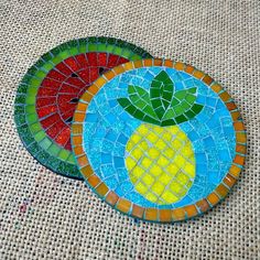 two pieces of colorful mosaic glass sitting on top of a cloth covered tablecloth, each with a pineapple in the center