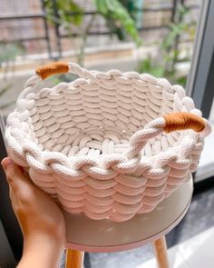 a person holding up a knitted basket on top of a stool next to a window