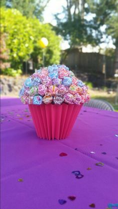 a pink cupcake covered in candy sitting on top of a purple table cloth with confetti sprinkles