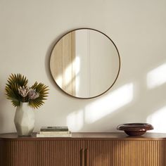 a round mirror on the wall above a wooden cabinet and vase with flowers in it