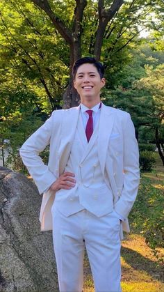 a man in a white suit and red tie standing next to a large rock with trees behind him