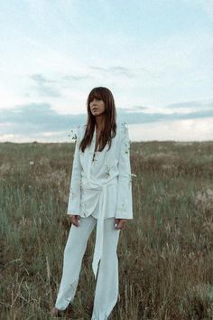 a woman standing in the middle of a field wearing a white suit with gold buttons