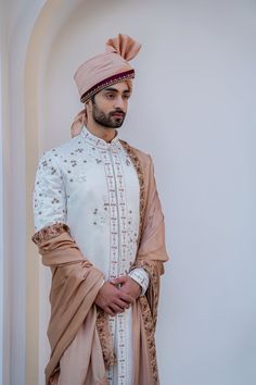 a man in a white and beige outfit standing next to a wall wearing a pink turban