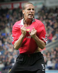 a man in red shirt and black shorts standing on soccer field with his hands together