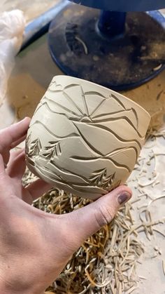 a hand holding a ceramic vase in front of a blue object on top of wood shavings