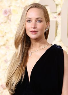 a woman with long blonde hair wearing a black dress and diamond necklace, standing in front of a floral wall