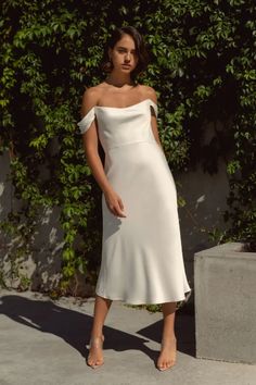 a woman wearing a white dress and sandals standing in front of a bush with her hands on her hips