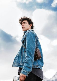 a young man wearing a jean jacket and holding a coffee cup in his hand while standing on top of a mountain