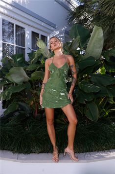 a woman in a short green dress standing next to some bushes and plants with her eyes closed