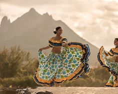 two women in colorful clothing are dancing on the beach with mountains in the background and clouds in the sky