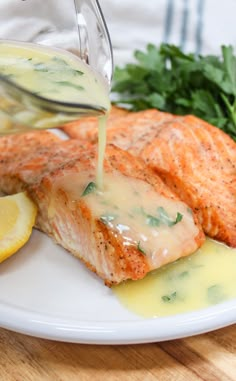 salmon being drizzled with gravy on a white plate next to lemon wedges and parsley