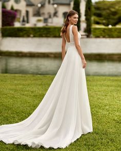 a woman in a white wedding dress standing on the grass near a lake and house