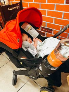 a baby sitting in a stroller next to a red brick wall and orange juice