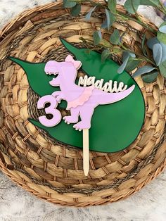 a green and pink dinosaur cake topper with the number three on it sitting in a wicker basket