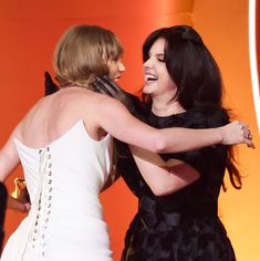 two women hugging each other on stage with an orange background and one woman wearing a white dress