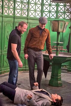 two men standing over a man laying on the ground in front of a green wall