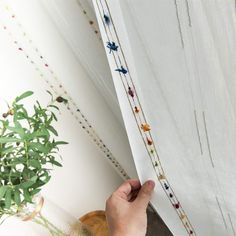 a hand is holding the edge of a curtain with beading on it and a potted plant next to it