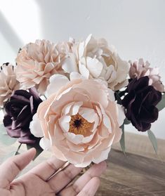 a hand holding a bunch of flowers on top of a wooden table in front of a white wall