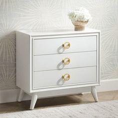 a white dresser with three drawers and two gold pulls on the handles, in front of a wallpapered background