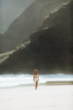 a woman is walking on the beach with her surfboard in hand and mountains in the background