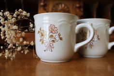 two white coffee mugs sitting on top of a wooden table next to some flowers