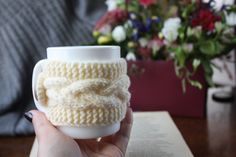 a hand holding a cup with a knitted design on it and a book in the background