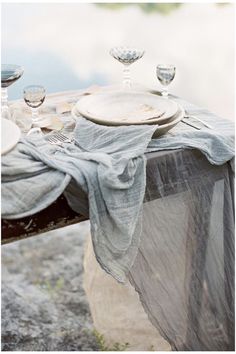 a table set with plates and silverware on top of a wooden bench next to water