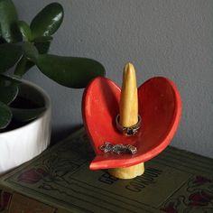 a red heart shaped container with a ring in it on top of a table next to a potted plant