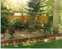 a wooden deck surrounded by plants and trees in a backyard with a bench on it