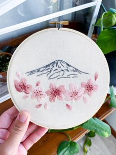 someone is holding up a embroidery project with pink flowers and a mountain in the background