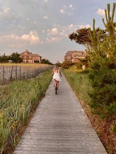 a woman riding a bike down a wooden walkway next to tall grass and houses in the background