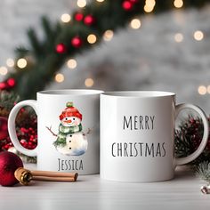 two white coffee mugs with merry christmas designs on them next to cinnamon sticks and decorations