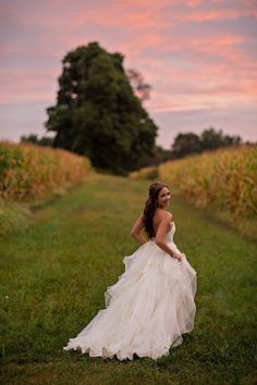 a woman in a wedding dress is standing on the grass near cornfield at sunset