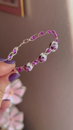 a hand holding a purple and white beaded bracelet with flowers on the side, in front of a pink wall