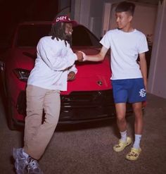 two young men shaking hands in front of a red sports car with the hood up