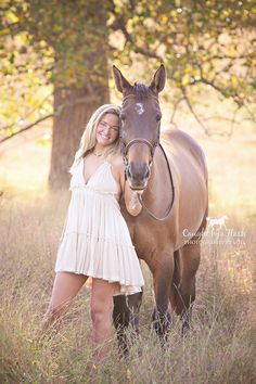 a beautiful blonde woman standing next to a brown horse in a field with tall grass