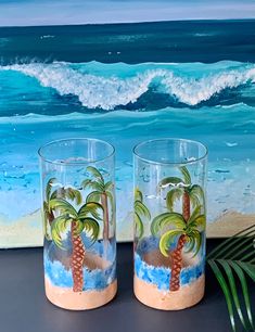 two glasses with palm trees painted on them sitting next to a painting of the ocean