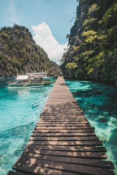 a long wooden dock leading to boats in the blue water near some mountains and trees