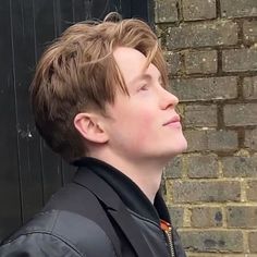 a young man standing in front of a brick wall wearing a black jacket and tie