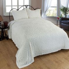 a bed with white bedspread and pillows in front of a window, next to a chair