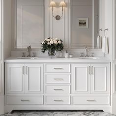 a white bathroom with two sinks, mirrors and flowers on the counter top in front of it