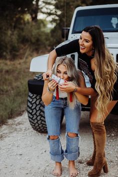 two women standing next to each other in front of a white truck and one is holding a camera