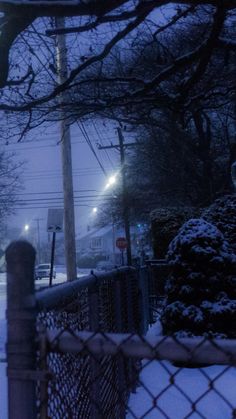 a snowy street at night with traffic lights on and trees in the foreground, behind a chain link fence