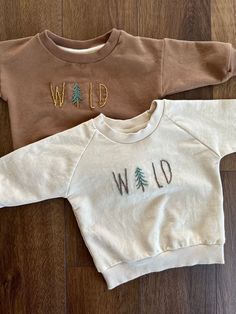 two sweaters that say wild and one with trees on the front, sitting on a wood floor