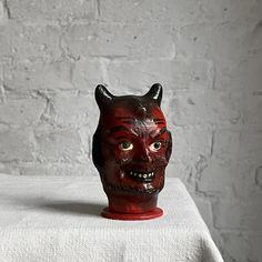 a red demon mask sitting on top of a white tablecloth next to a brick wall