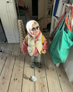 a small child wearing sunglasses and a scarf standing in front of a door with bags on the floor